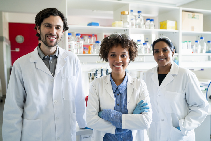 Portrait of young scientist standing arms crossed with colleagues. Smiling male and female researchers are working together in laboratory. They are wearing lab coats.