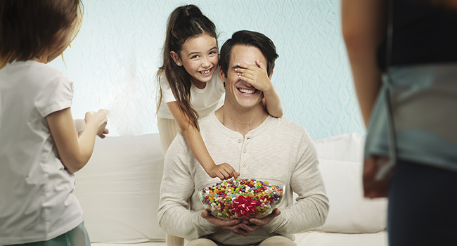 A little girl covering her Dad's eyes while she grabs a handful of jelly beans from a bowl he is holding while his other daughter and wife watch