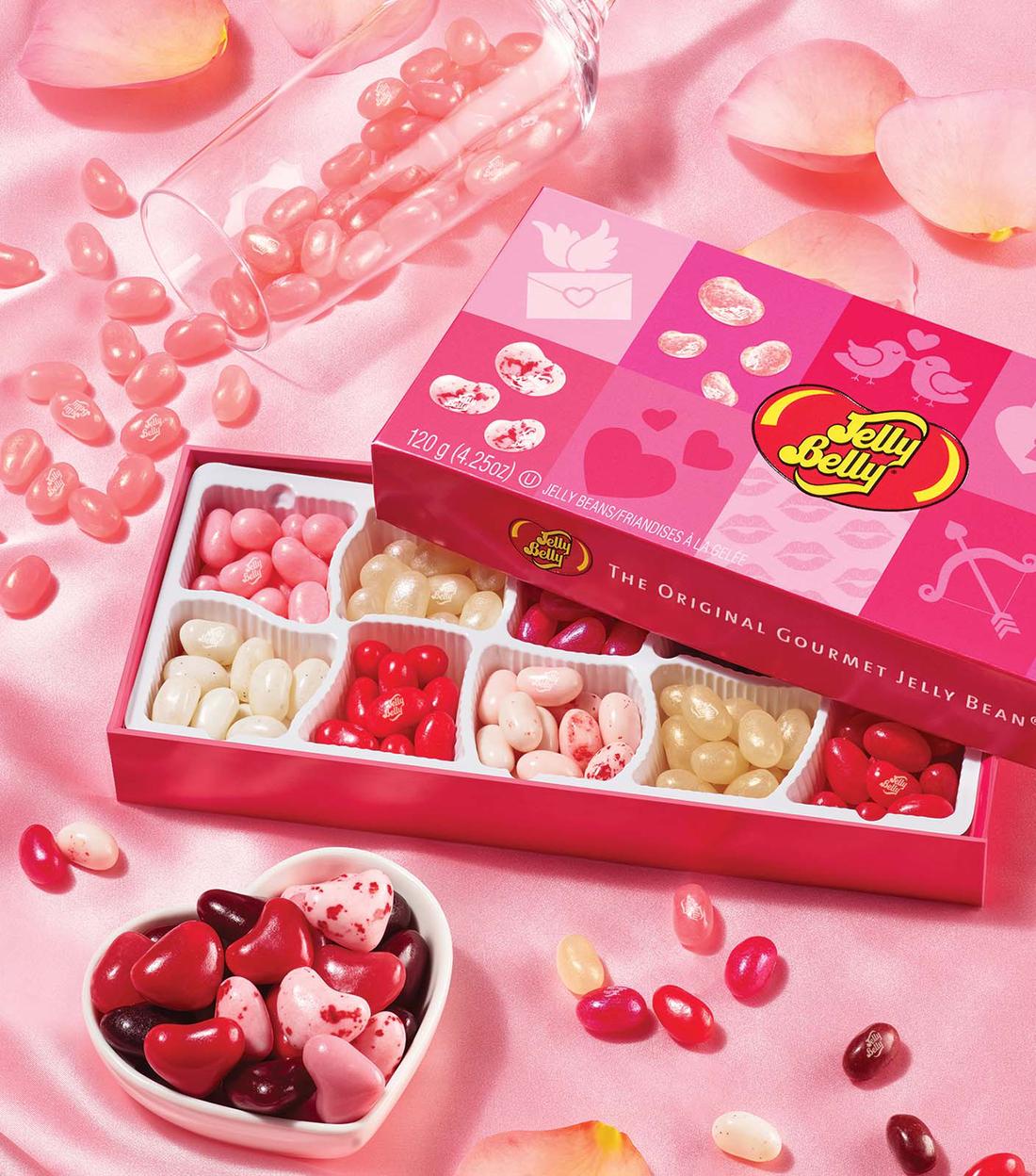 A valentines box of jelly beans next to a wine glass of spilled pink jelly beans and a heart shaped bowl filled with heart shaped candy